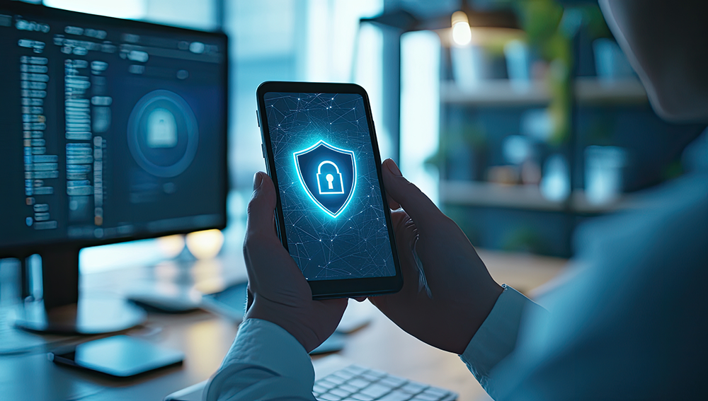 Person holding a smartphone displaying a glowing security shield icon, symbolizing mobile cybersecurity, with computer monitors in the background.